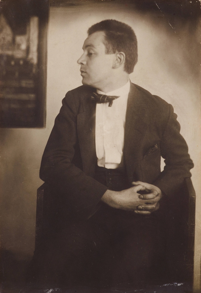 Photograph of Egon Schiele in profile with clasped hands, in front of his painting Houses by the Sea, taken by Anton Josef Trčka in 1914. Leopold Museum, Vienna.