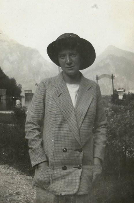 With Arthur Roessler, Walburga Wally Neuzil during an excursion, by Arthur Roessler, circa 1913-1914. A vintage black-and-white photograph showing Wally Neuzil outdoors, facing forward with her hands in the pockets of a double-breasted checkered jacket, wearing a wide-brimmed hat, with Austrian mountains visible in the hazy background.