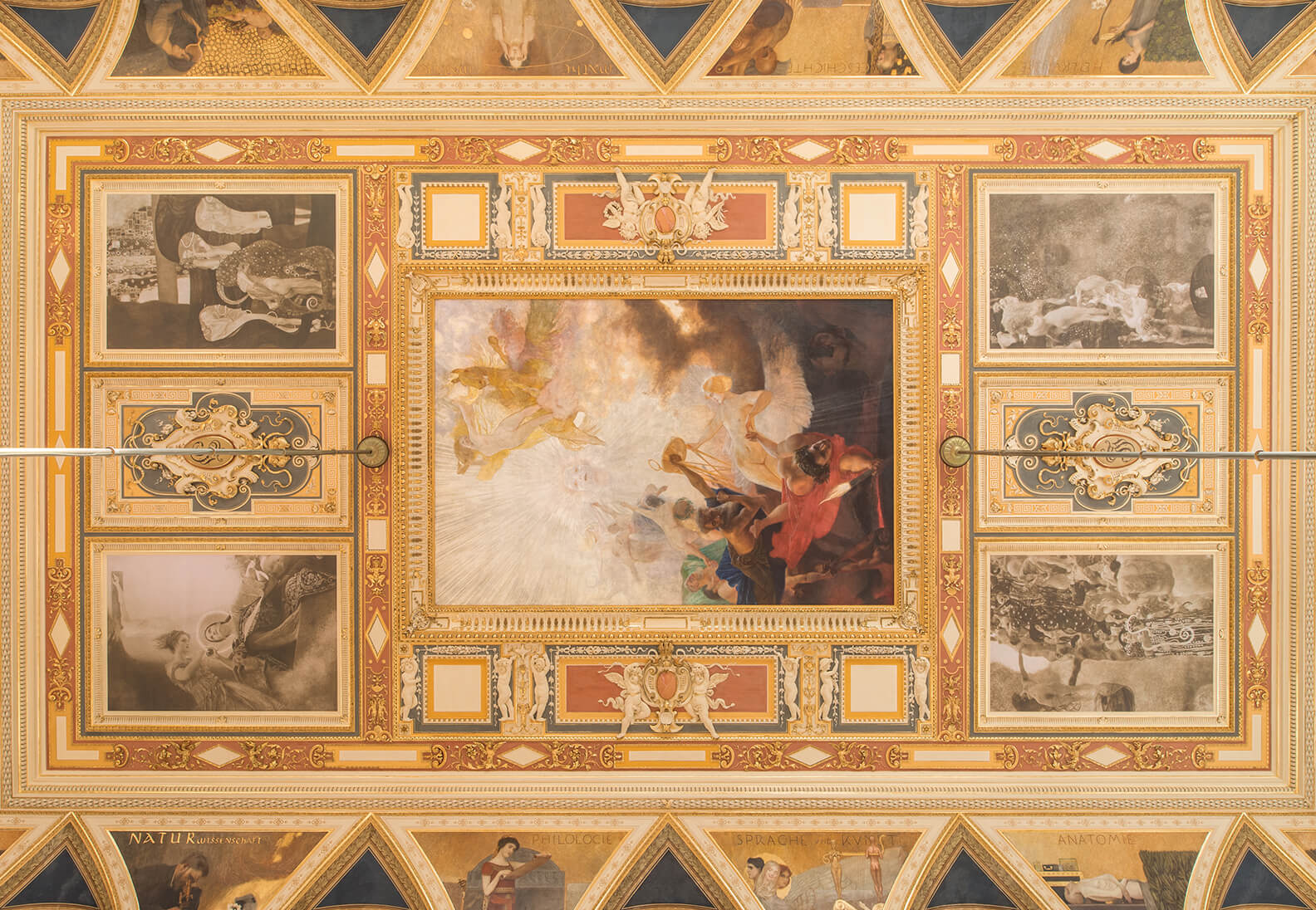 Digital photo of an architectural ceiling reconstruction installation, Planned Arrangement of the Faculty Paintings in the Great Hall of the University of Vienna, by Gustav Klimt and Franz Matsch, 2019. University of Vienna. A full view of an ornate, gilded stucco ceiling structure containing five decorative panels arranged around a larger central painting.