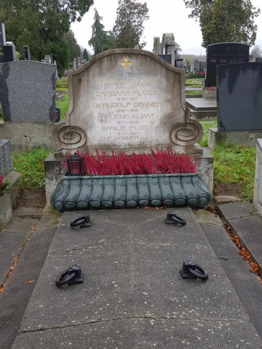 Photograph, The grave of the Flöge and Klimt family at the Evangelischer Friedhof Simmering, by Schiele & Klimt, 2025. Front view of a large stone family grave monument with a curved top and decorative volutes, listing the names and dates of Pauline Flöge, Barbara Flöge, Dr. Rudolf Donner, Helene Klimt, Emilie Flöge, and Helene Donner geb. Klimt. A planter with red heather flowers sits in front of the headstone.